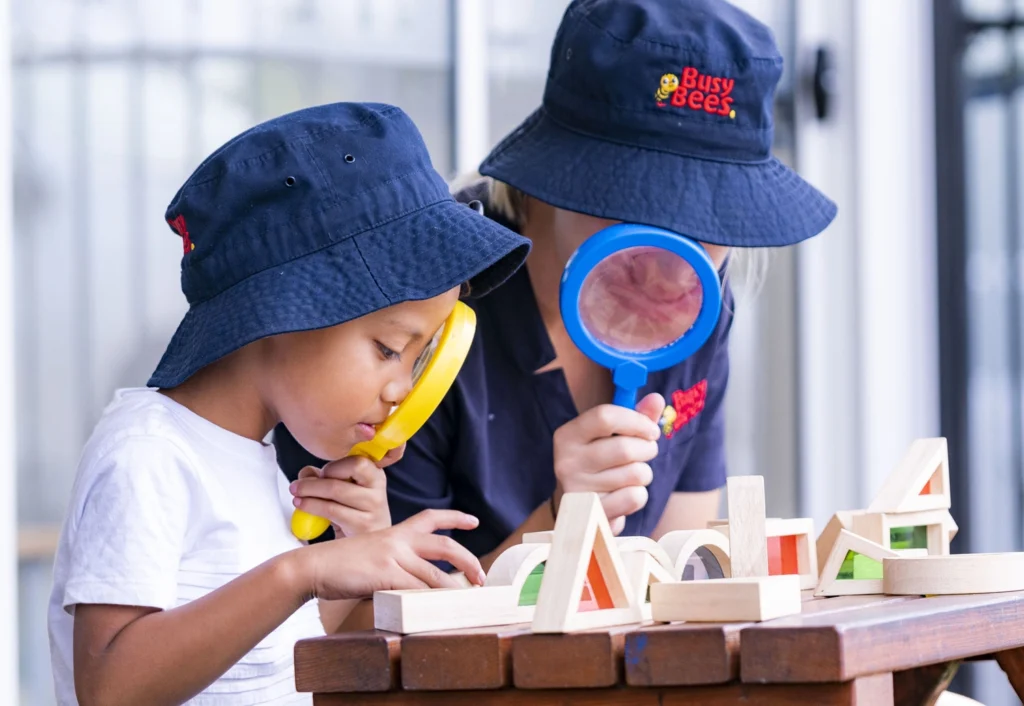 Two children wearing navy blue hats examine wooden blocks on a table with magnifying glasses.