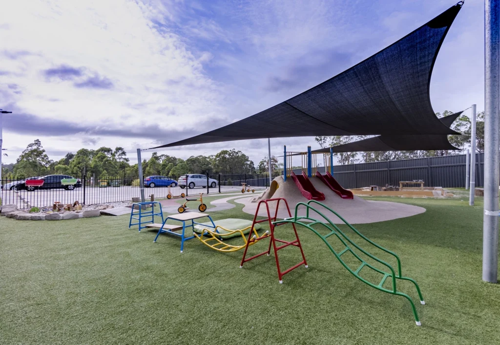 A children's playground with climbing equipment, slides, and tricycles on an artificial turf surface. A large shade sail covers part of the area. Cars and trees are visible in the background.