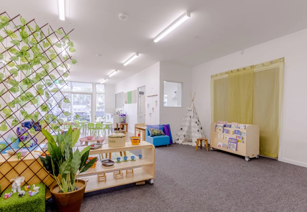 A well-lit classroom with plants, a teepee, bookshelves, educational toys, and a large window.