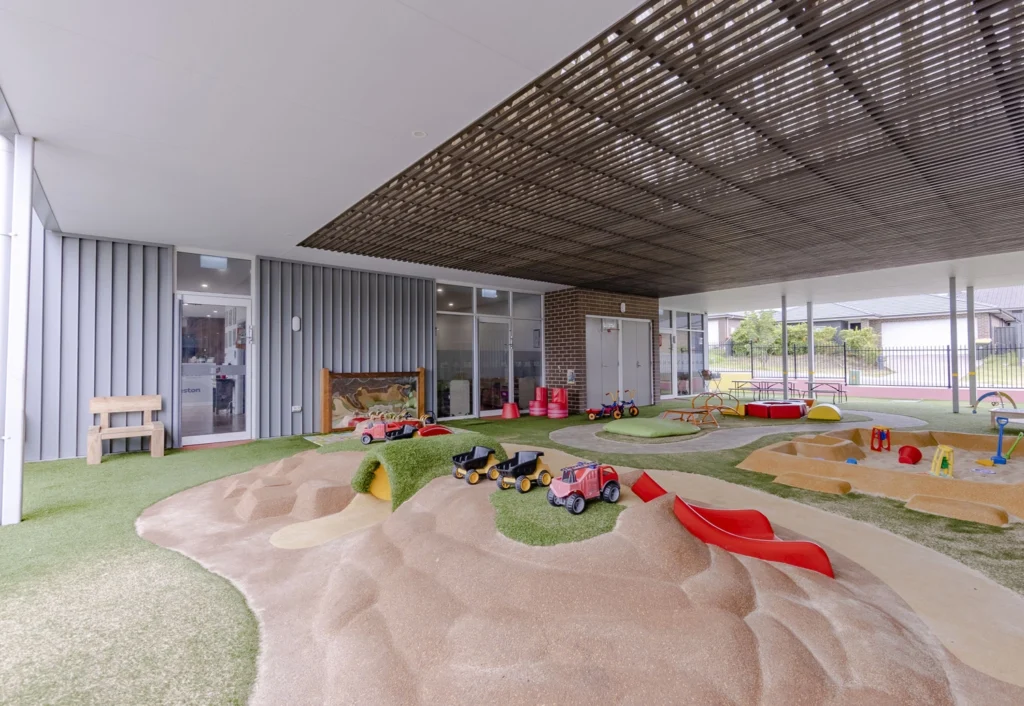 A children's play area with toy trucks, slides, and a sandbox under a shaded canopy with artificial turf flooring.