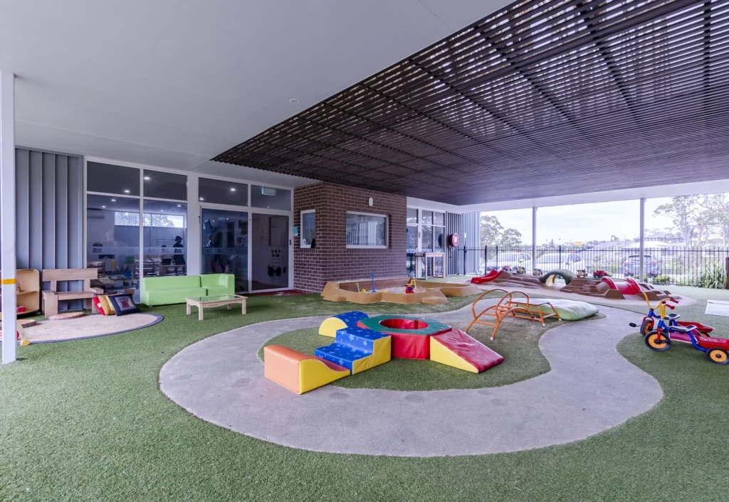 A covered outdoor play area with various children's play equipment including a slide, climbing structures, a sandpit, toy vehicles, and seating. The ground is covered in artificial grass.