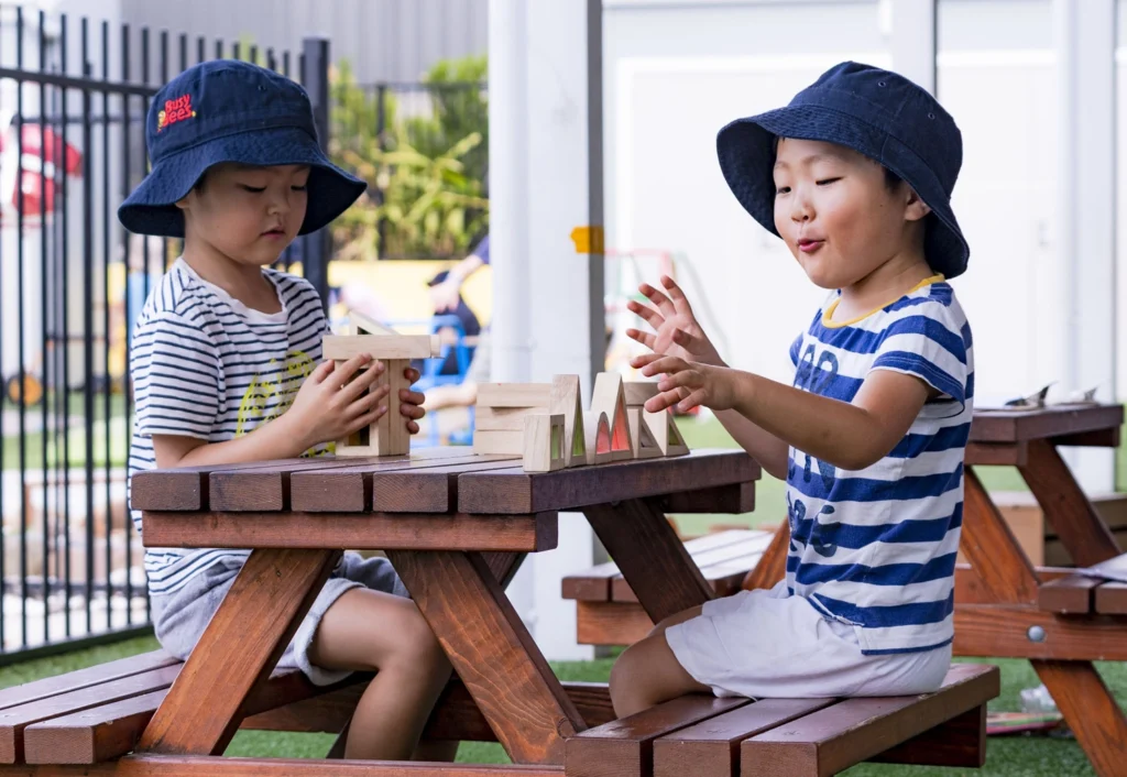 Two children wearing sun hats sit at a wooden picnic table playing with wooden blocks. They are outdoors on a sunny day.