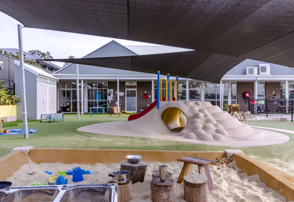 A playground with a slide, tunnel, and climbing mound under shade. The area also has a sandbox with toys and stumps for seating. Buildings and equipment are visible in the background.