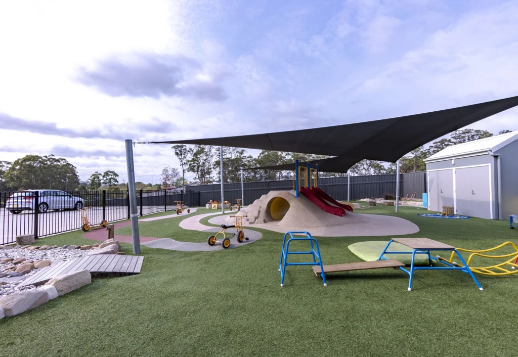 Outdoor playground with artificial grass, climbing equipment, tricycles, a slide, and a sandpit cover. A shaded area provides protection, and a fence encloses the playground.