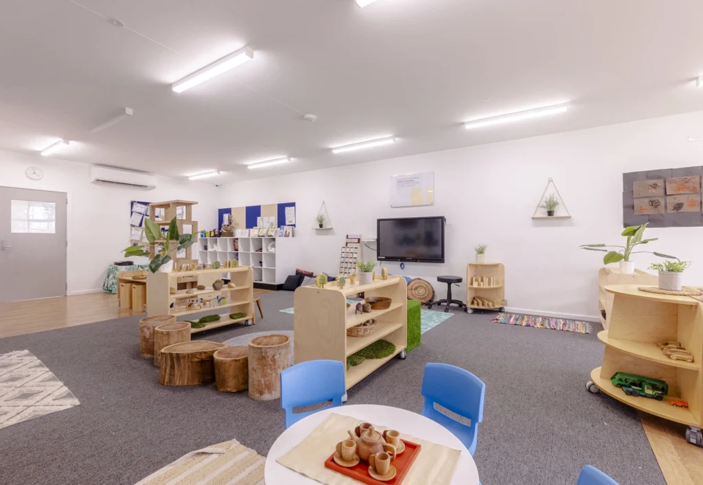 A spacious and well-lit classroom with child-sized furniture, wooden play structures, indoor plants, and learning materials neatly arranged. A large screen is mounted on the wall at the front.