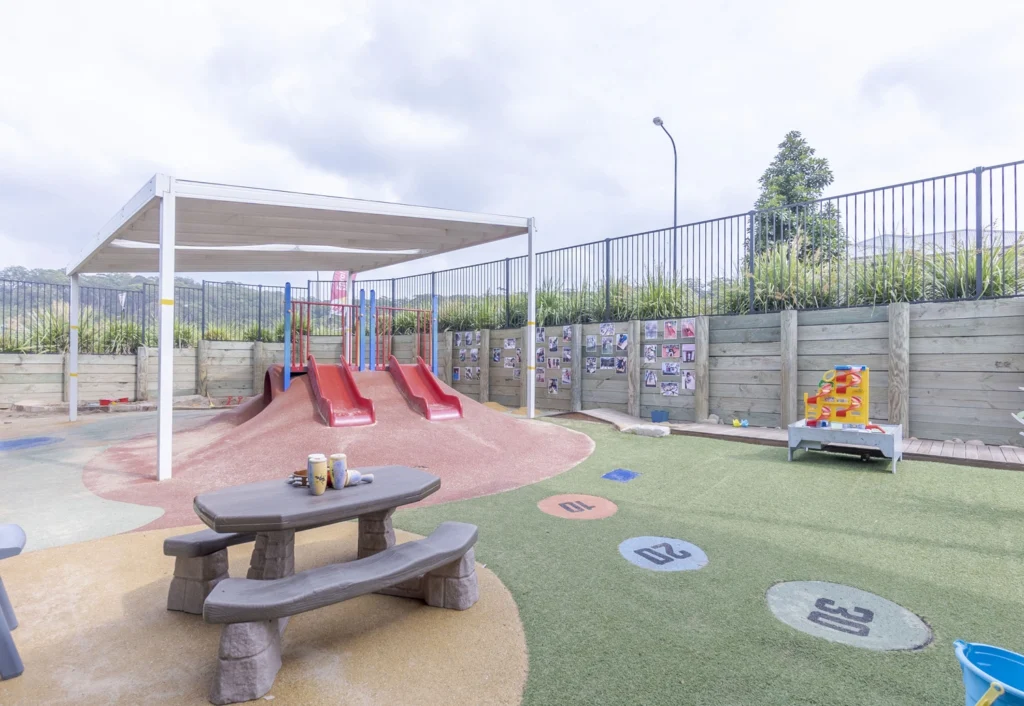 Outdoor playground with red slides, a covered picnic table, and hopscotch painted on the ground. A wooden fence with pictures and a toy table are visible in the background.