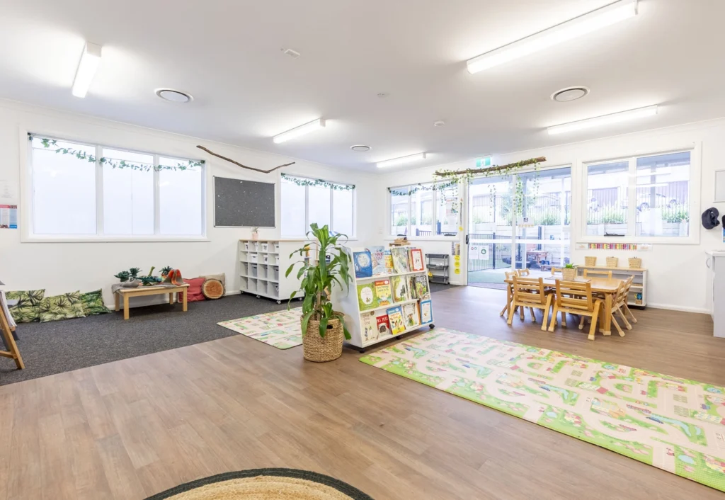 A brightly lit, spacious classroom with a reading area, small tables and chairs, a whiteboard, bookshelves, and plants. Large windows and a glass door allow natural light to fill the room.