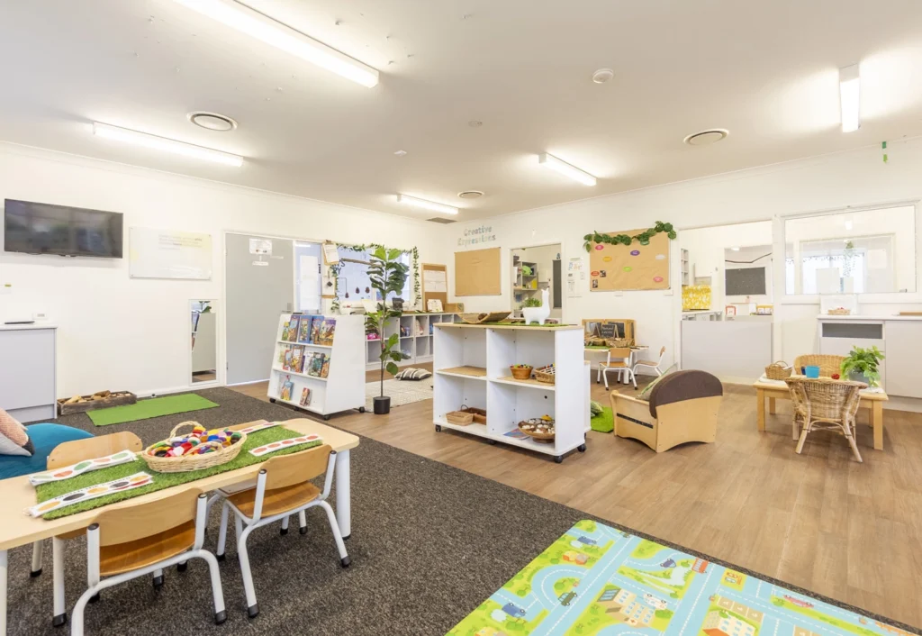 A bright classroom with tables, chairs, play mats, bookshelves stocked with books, a flat-screen TV on the wall, and various educational materials and toys organized throughout the space.