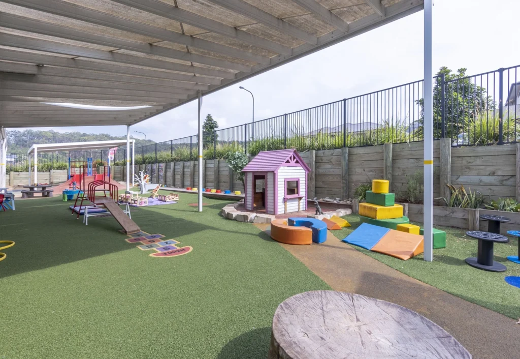 Outdoor play area featuring a small playhouse, hopscotch game, colorful foam blocks, and various playground equipment on artificial grass under a covered area.