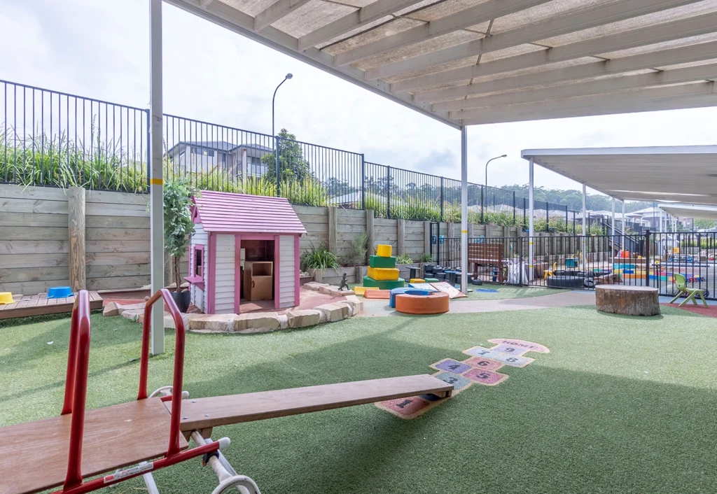 Outdoor playground area with a small pink playhouse, seesaw, and various climbing structures on an artificial grass surface. The space is fenced and covered by a canopy.