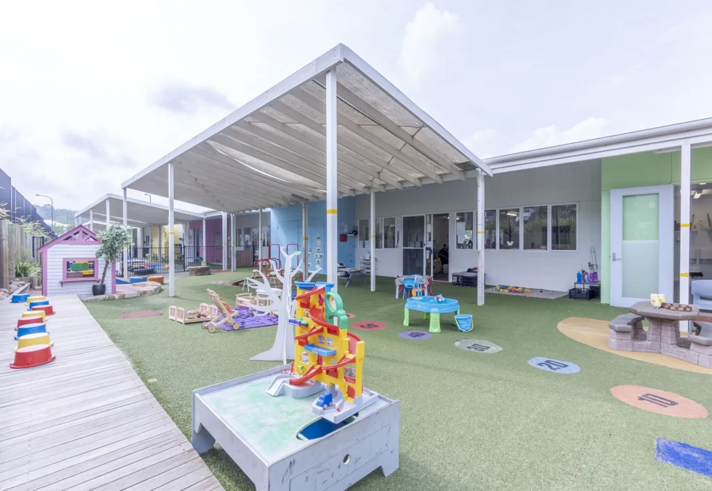 An outdoor children's play area with various colorful toys and play equipment, situated adjacent to a modern, white-roofed building.