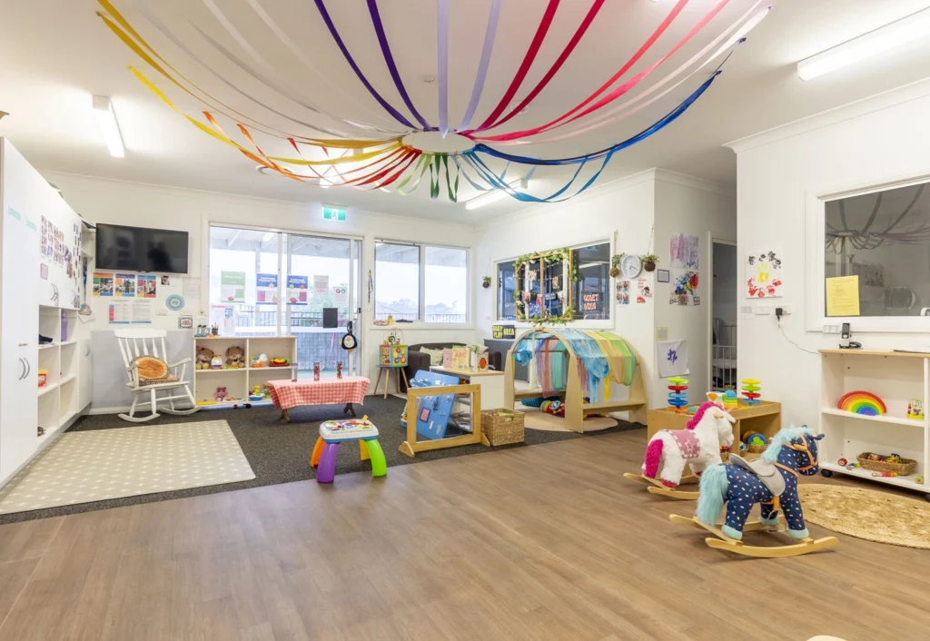 A brightly decorated nursery room with colorful streamers on the ceiling, various toys, a rocking chair, and play areas with activities for young children.