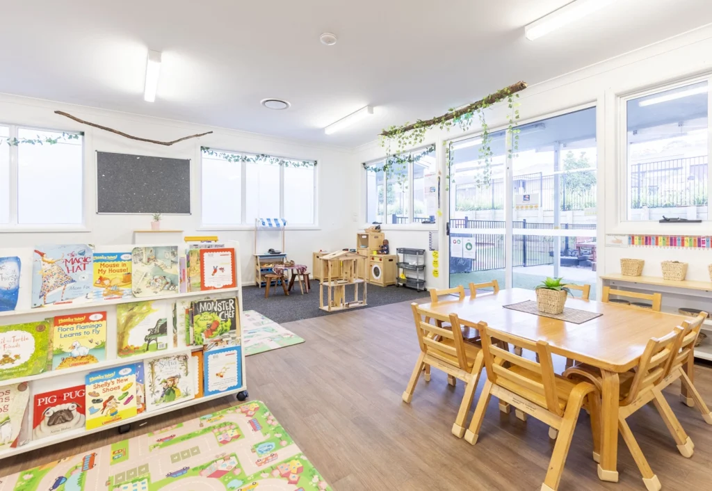 A brightly lit classroom with bookshelves, books, a reading mat, small wooden chairs and tables, and various learning materials. Large windows allow natural light, with plants hanging from the ceiling.