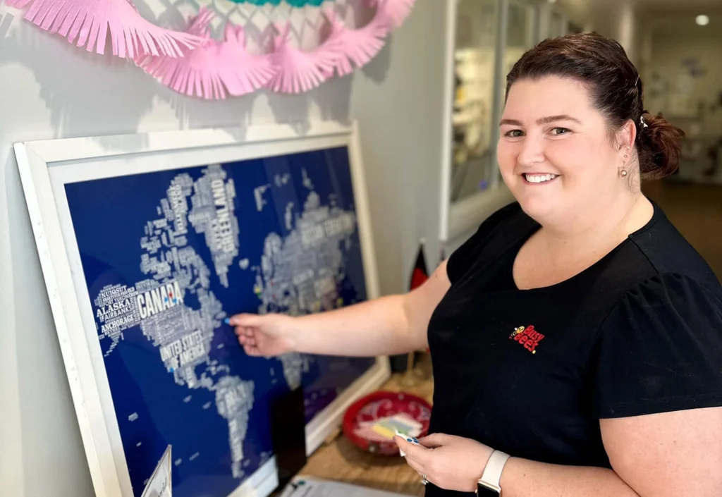 A woman stands indoors, smiling and pointing at a large world map display. The map highlights various countries with text. She wears a black shirt and a watch on her left wrist.