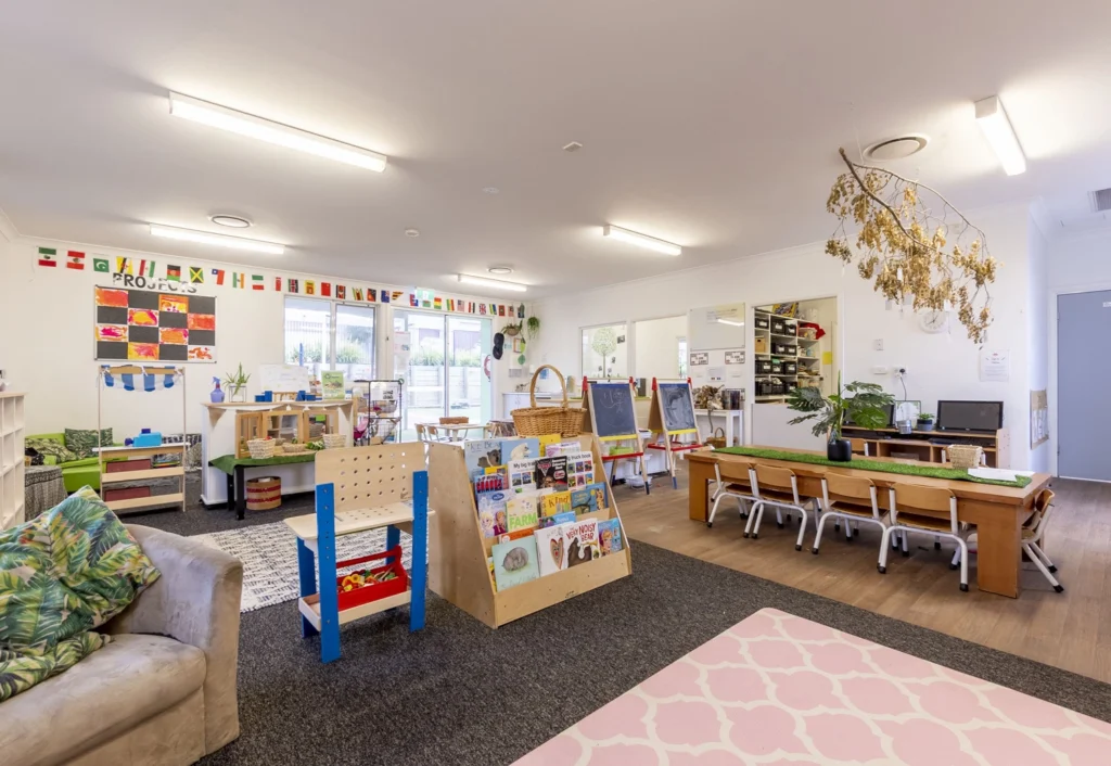 A brightly lit preschool classroom with various activity stations, including bookshelves, small tables with chairs, and colorful decorations on the walls.