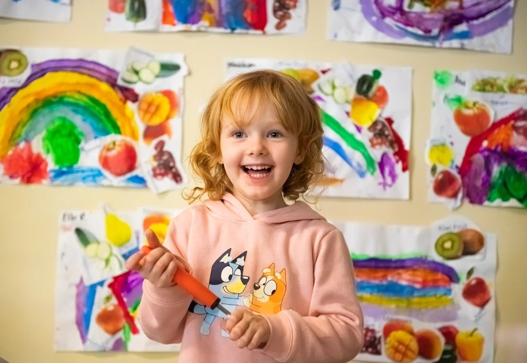 A Kindergarten girl with curly red hair smiles in front of a colorful wall displaying various children's artwork at a Cannington day care.