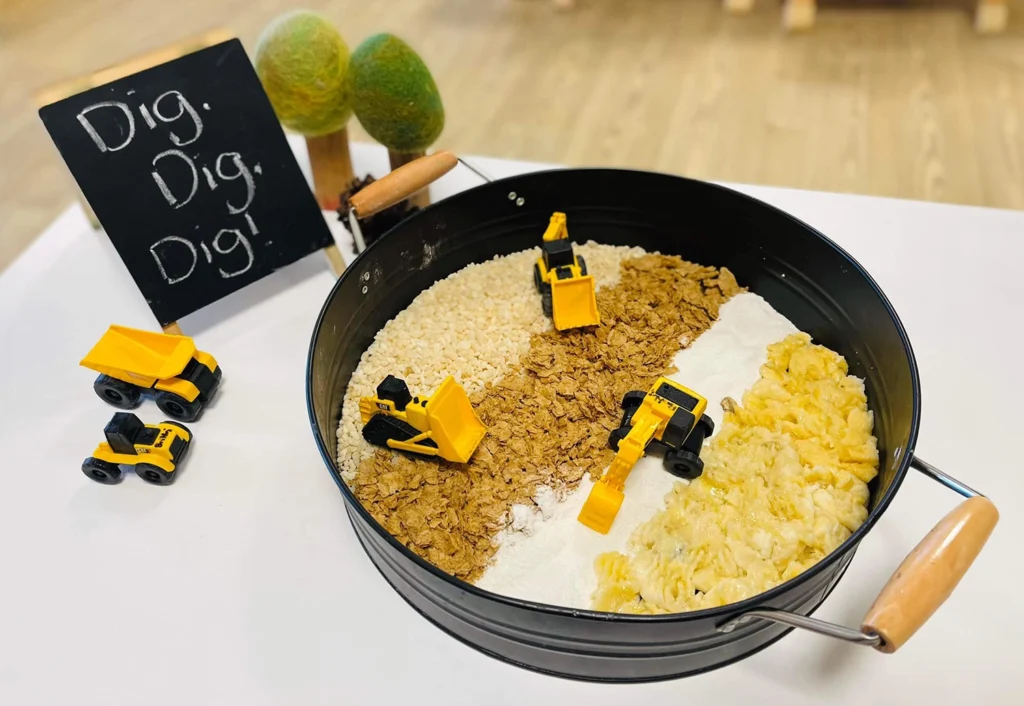 A black round tray contains separate sections of rice, cornflakes, flour, and pasta, each with small toy construction vehicles. A blackboard with "Dig, Dig, Dig!" written on it stands beside the tray.