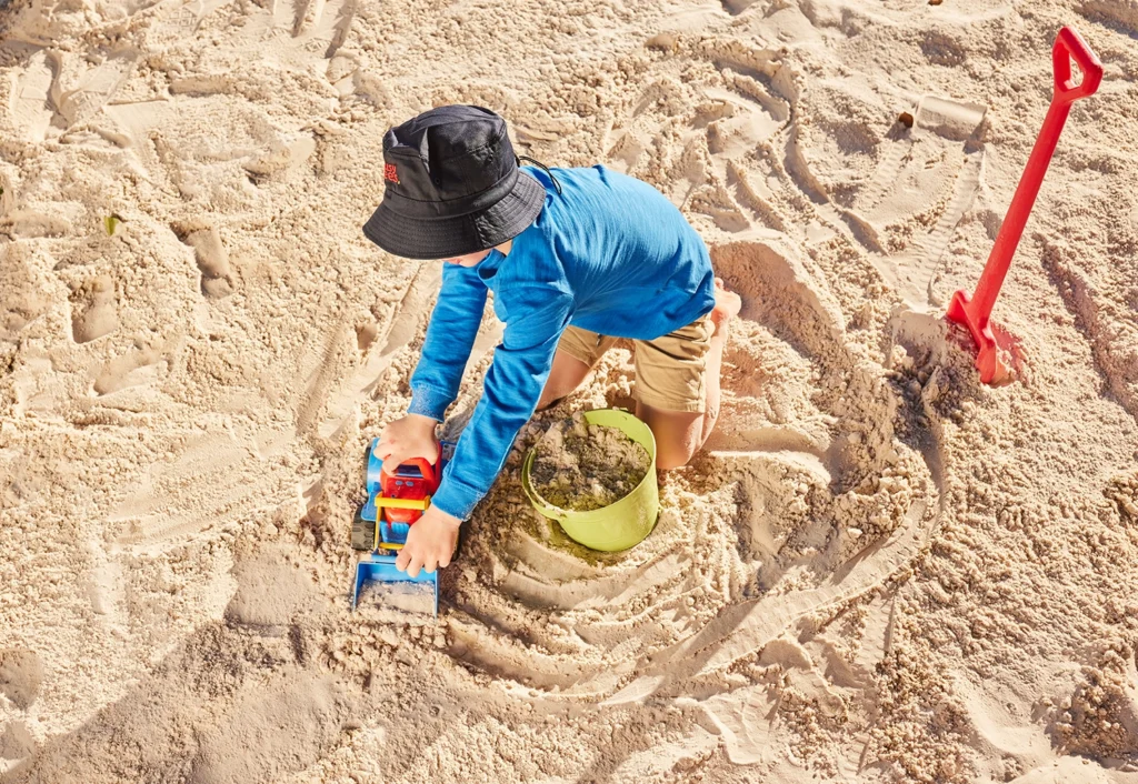 Child in a blue shirt and bucket hat plays with a toy bulldozer in the sandpit at Busy Bees Cannington childcare.