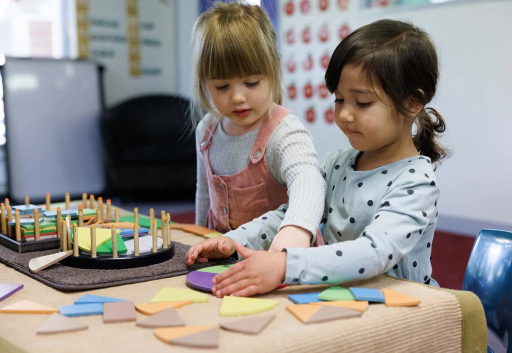 Two young girls are engaged in an indoor activity, working together with colorful geometric shapes on a table. One girl is reaching for a purple shape, while the other is assembling shapes in front of her.