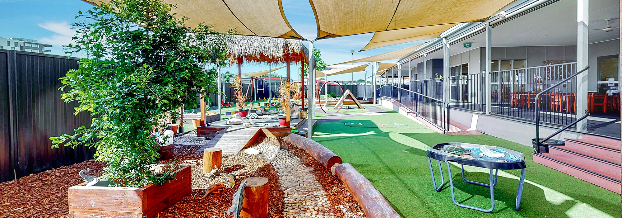 Outdoor children's play area with artificial grass, wooden logs, and shaded structures. There are various play equipment pieces and a small table. A building with large windows is adjacent to the play area.