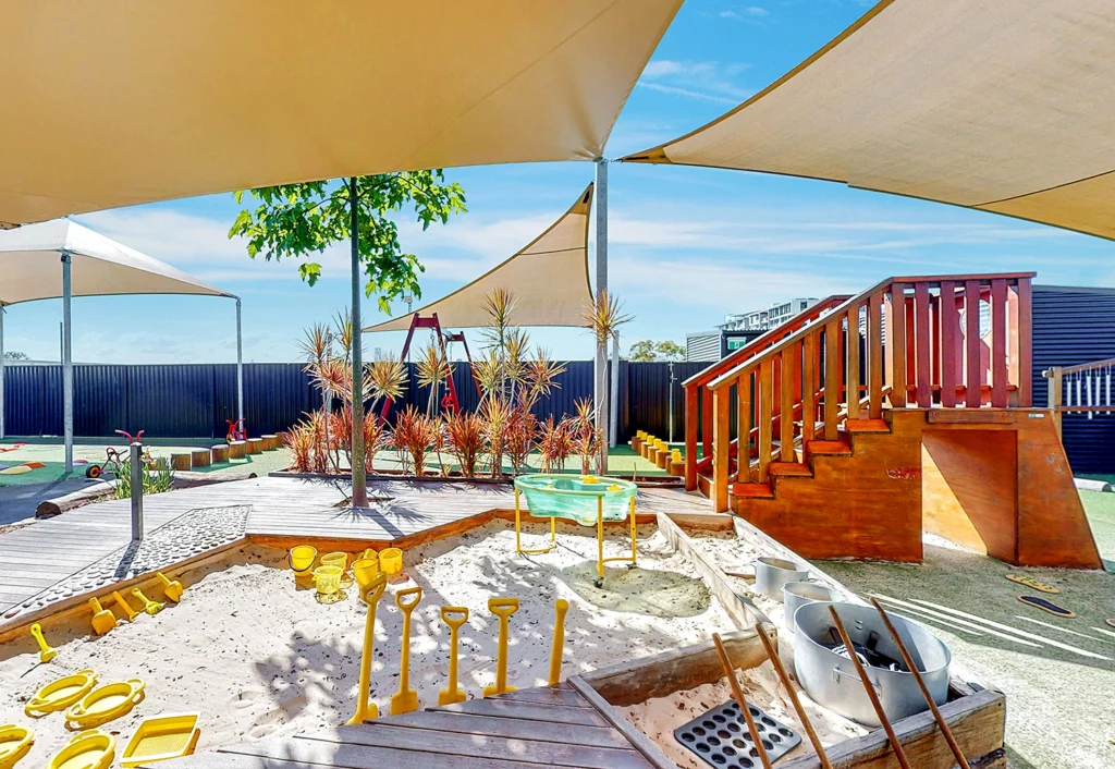 A children's playground with shade sails, sandpit, wooden steps, and yellow buckets and shovels. The background features plants and a black fence.