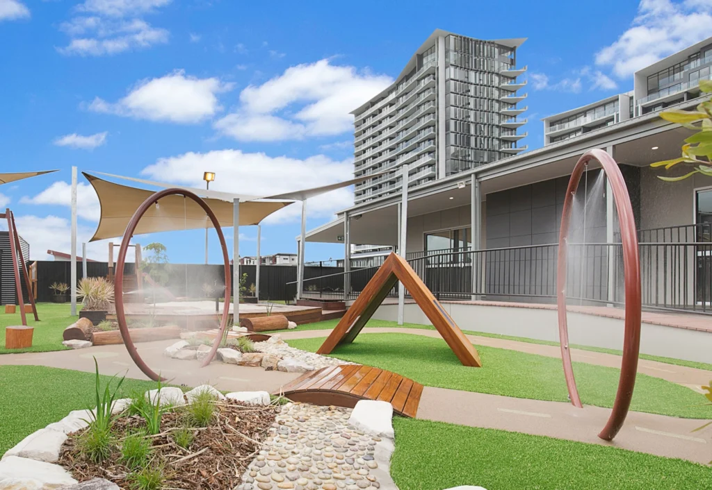 Modern outdoor playground with water play features, shaded seating areas, artificial turf, and landscaping. High-rise buildings are in the background under a blue sky with scattered clouds.