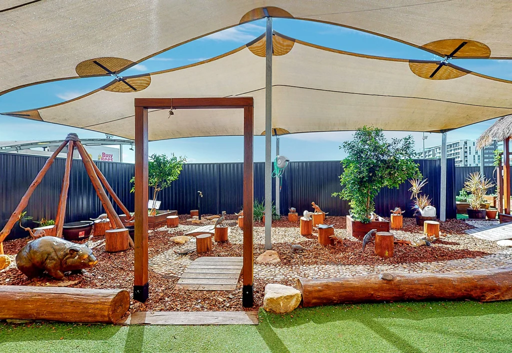 A playground area with wood structures, a small swing set, various plants, and a shade canopy overhead. The ground is covered with mulch and stepping stones. A wombat statue is placed near the entrance.