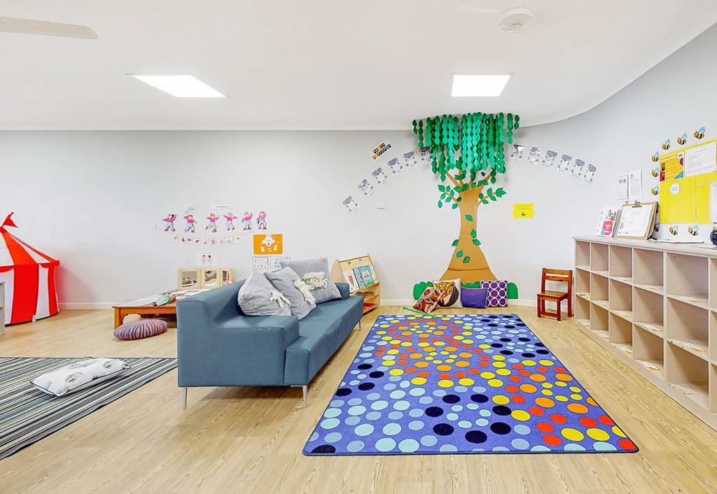 A brightly lit children’s playroom with a colorful carpet, a blue sofa, a red and white tent, wall decorations, and a tree mural. Shelves line one wall, holding various toys and books.