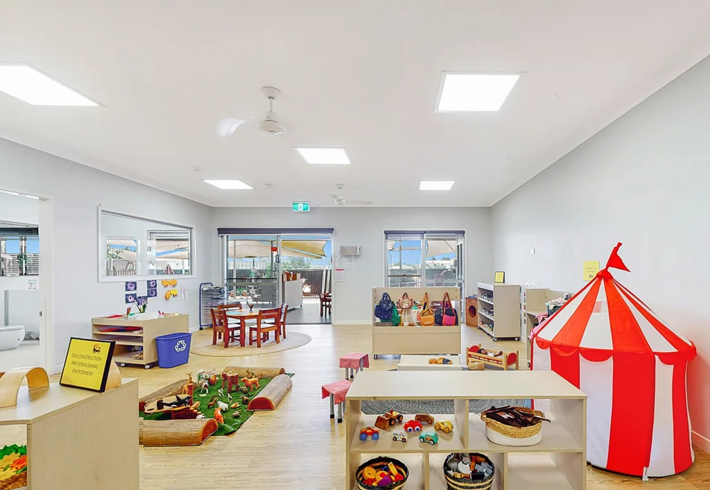 A well-lit children's playroom with various toys, a red and white play tent, small tables, and storage shelves. There are windows along the back wall providing natural light.