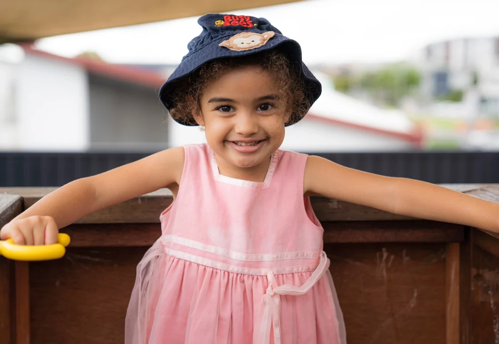 A young girl wearing a pink dress and a blue hat stands smiling with her arms outstretched, holding a yellow handle.