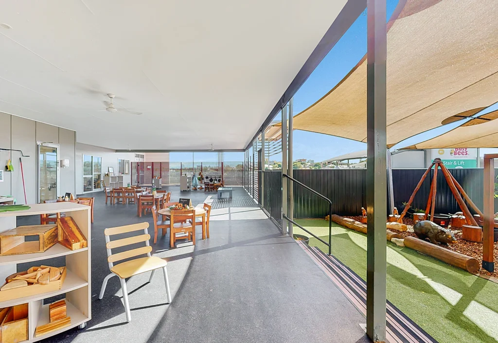 Interior view of a brightly lit room with children's furniture, toys, and play area, adjoining an outdoor playground with shade sails overhead.