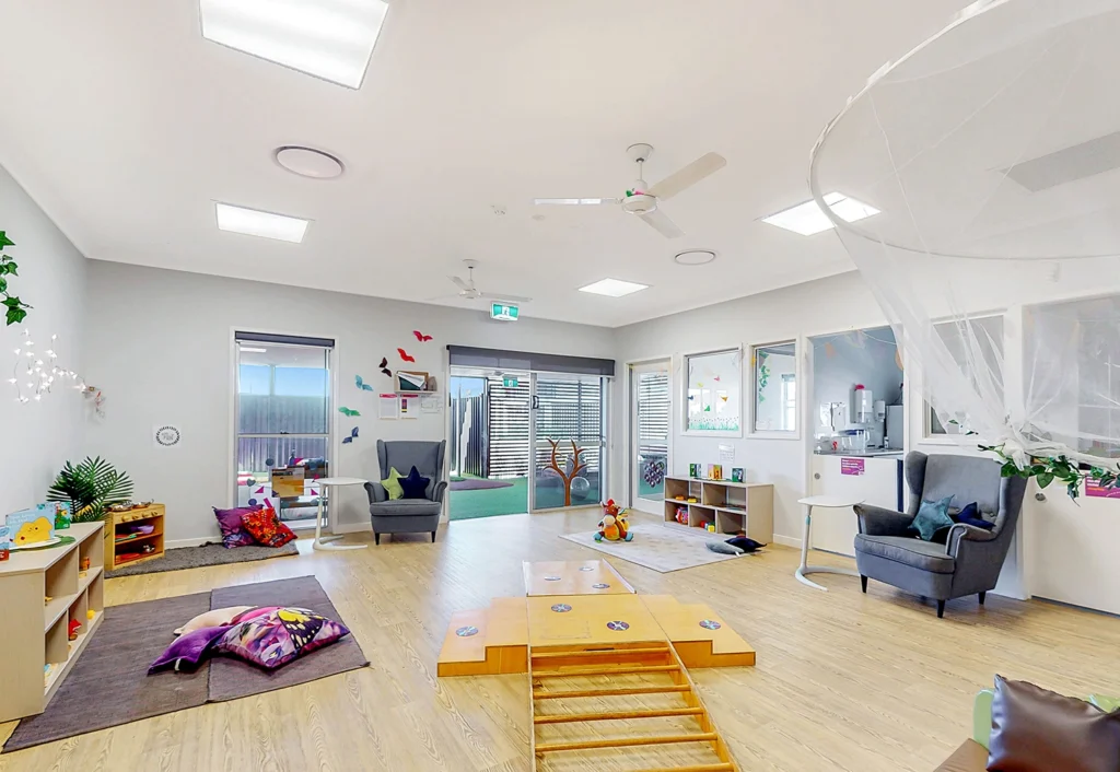 A spacious, well-lit room with children's play area, wooden shelves, plush seating, toys, and craft items. The room features large windows, a play structure, and a mesh canopy over an armchair.