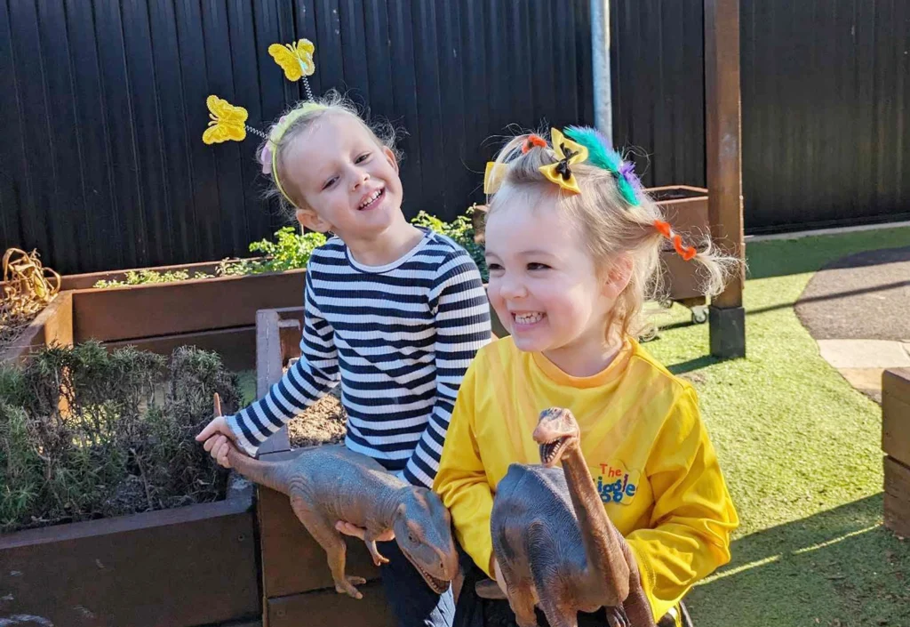 Two children smiling outdoors, holding toy dinosaurs. One child is dressed in a striped shirt with butterfly hair clips, and the other wears a yellow shirt with colorful hair ties.