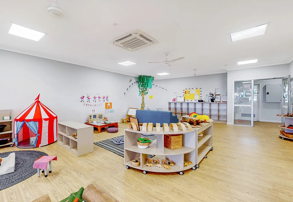 A spacious, well-lit children's playroom with various toys, a small play tent, low shelves holding books and baskets, colorful wall decorations, and wooden flooring.