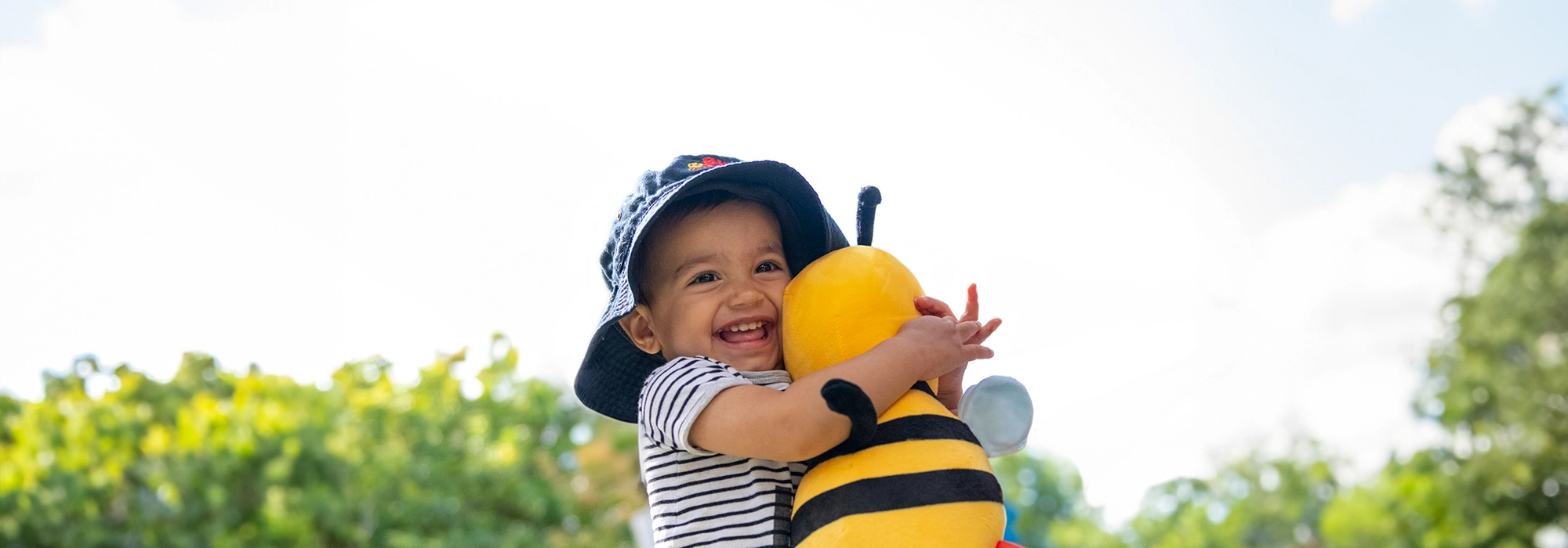 A smiling child in a striped shirt and bucket hat hugs a plush toy bee outdoors with greenery and a clear sky in the background.