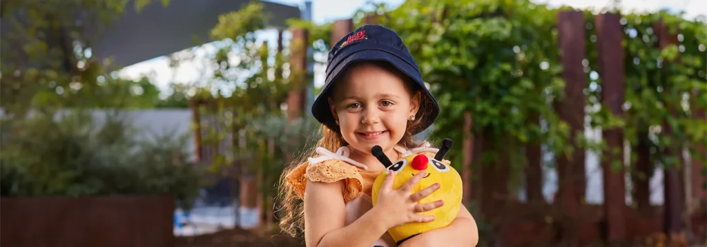 A young girl wearing a navy blue hat smiles while holding a yellow stuffed toy in a sunny outdoor setting with greenery in the background, capturing the joy of her preschool adventures.