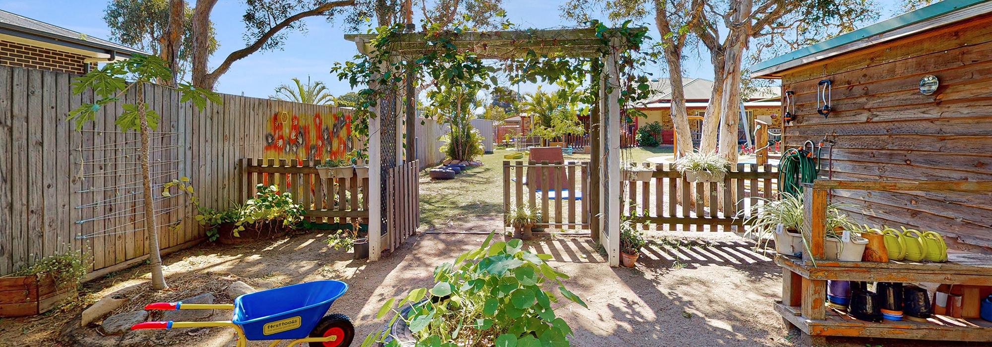 "Garden World" at Busy Bees Hervey Bay day care with a wooden arch entrance, into a fenced area with vegetable and herb gardens and a wheelbarrow.