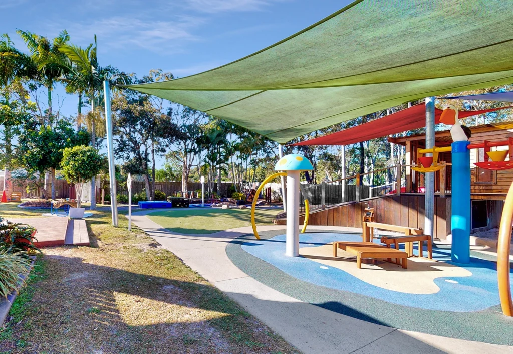 A children's outdoor playground at Hervey Bay Childcare featuring a colorful water play area with tipping buckets, water misters and a hand water pump. In the background is a timber fort and spacious green lawn areas surrounded by trees.