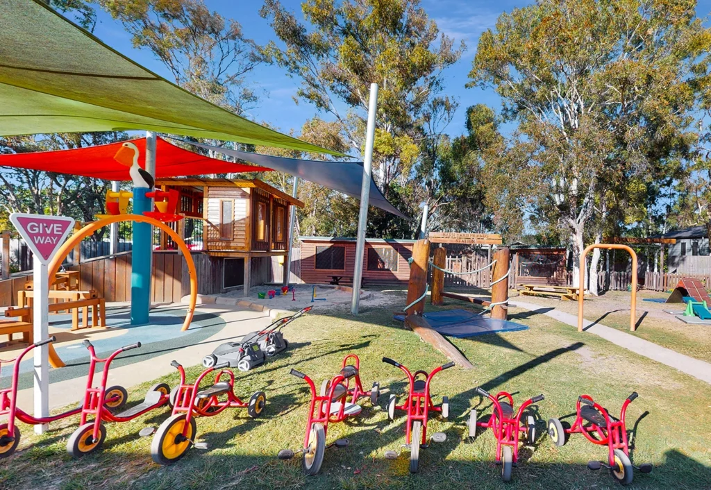 A children's playground at a day care in Hervey bay with red tricycles lined up on grass, play structures, a sandpit and trees in the background.