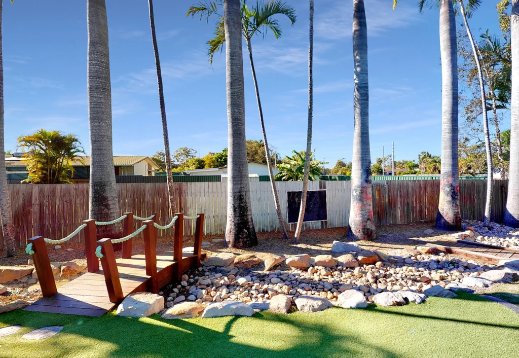 The outdoor play space at Hervey Bay Childcare centre featuring a wooden bridge crossing a dry creek bed surrounded by tall trees and green lawn.