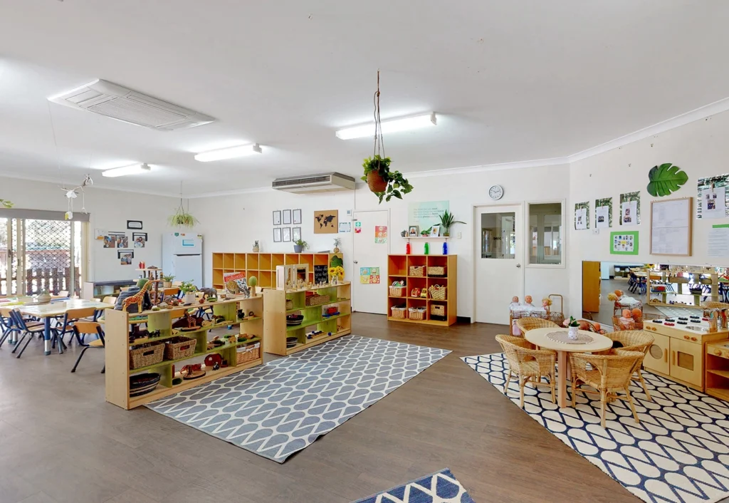 A brightly lit kindergarten classroom with wooden furniture, educational toys, shelves, and tables. The room has plants, rugs, and wall decorations, creating a welcoming early learning environment at Hervey Bay Day Care.