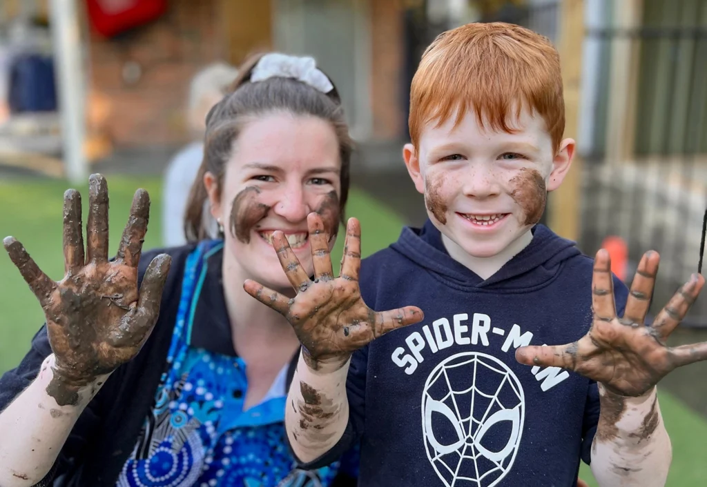 A early learning educator and kindergarten boy smile at the camera showing their muddy hands and faces after a messy play experience at Busy Bees Fraser Coast located in Hervey Bay