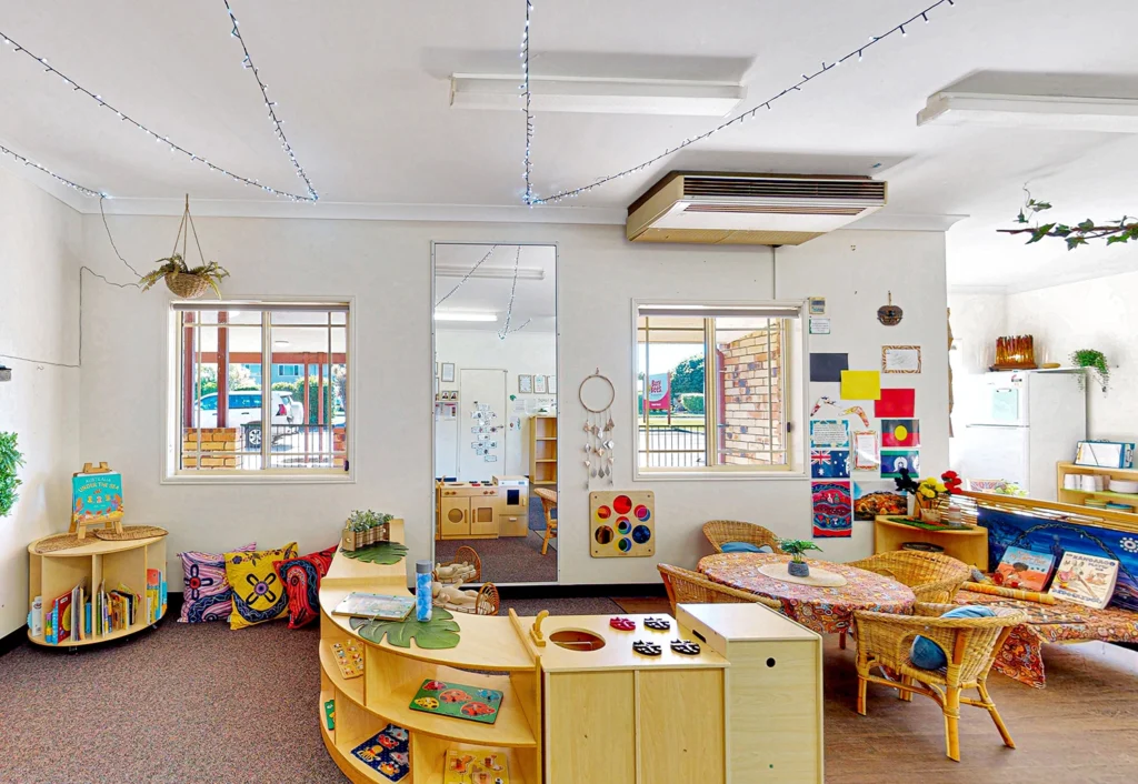 A brightly lit classroom at Busy Bees Hervey Bay featuring colorful bookshelves, woven chairs, and a variety of toys and educational materials.