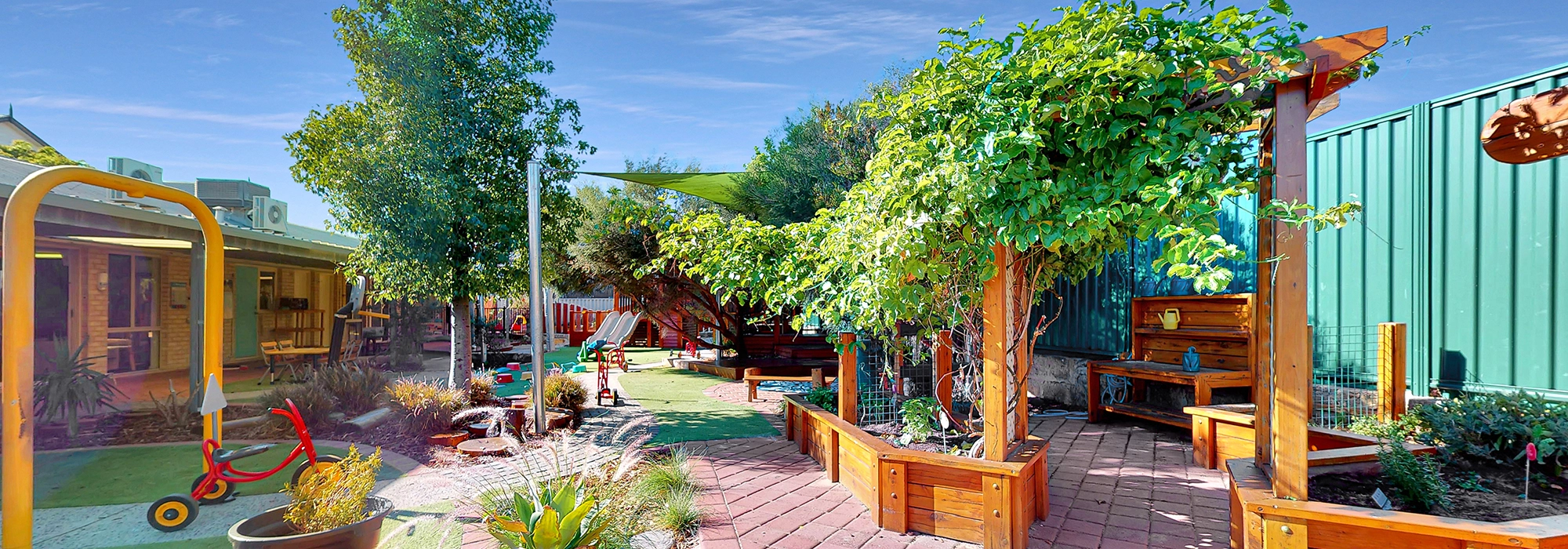 A sunny, well-landscaped garden with wooden planters, green shrubs, a tricycle, and various children's play equipment beside a building and a green metal fence.