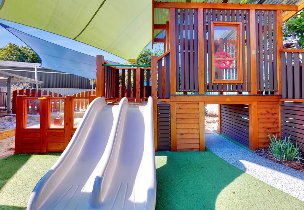 A children's outdoor play area featuring a double slide and an elevated wooden playhouse with a canopy providing shade. The ground is covered with artificial grass.