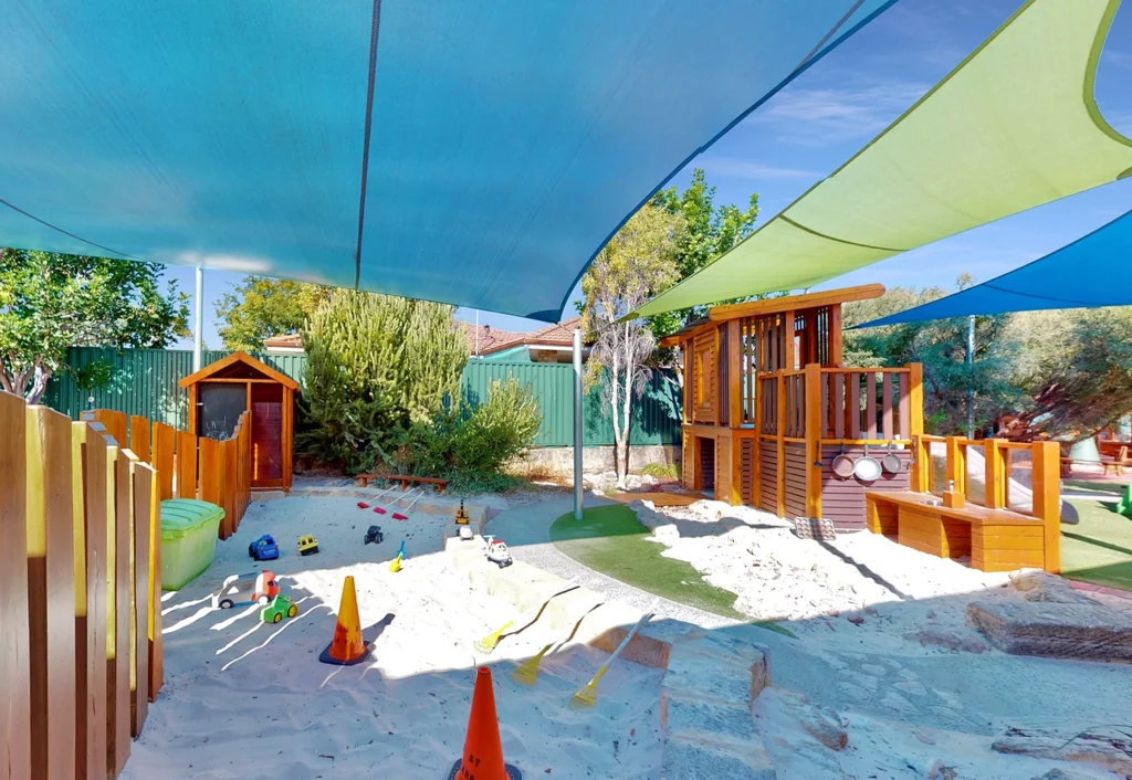 A playground featuring a sandbox with various toys, wooden play structures, and shade sails overhead for protection from the sun. A small wooden playhouse is visible in the background.