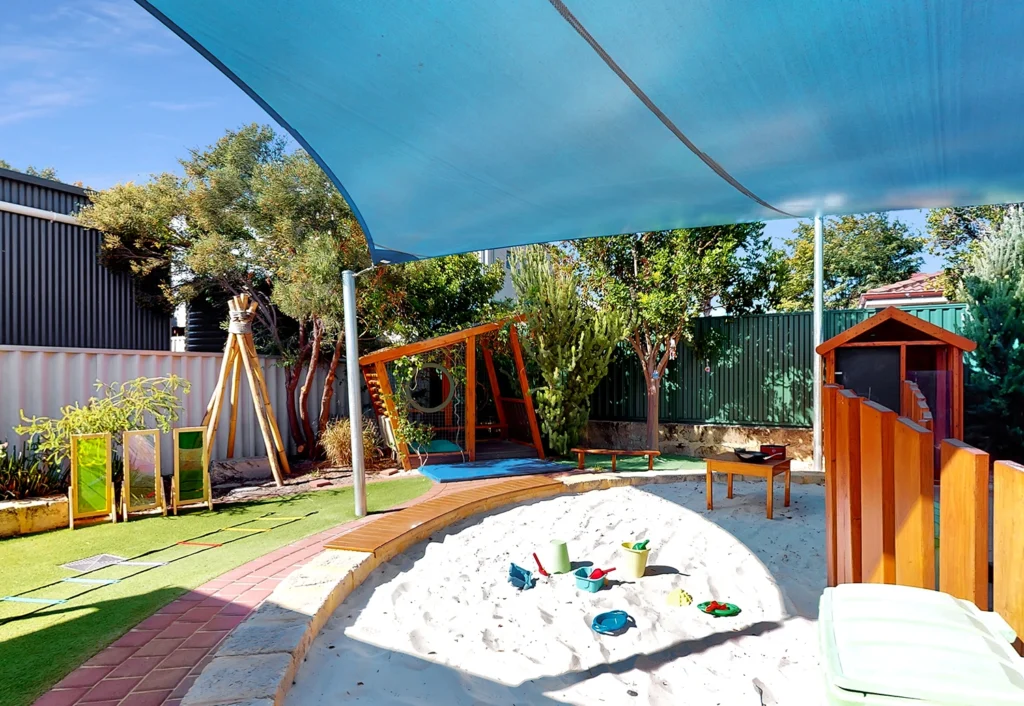 A playground area with a sandbox, toys, shaded seating, and play structures. There is a blue shade canopy overhead and greenery surrounding the space.