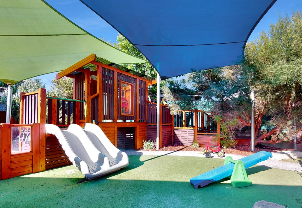 A children's playground features a wooden play structure, slides, and seesaws, shaded by fabric canopies.