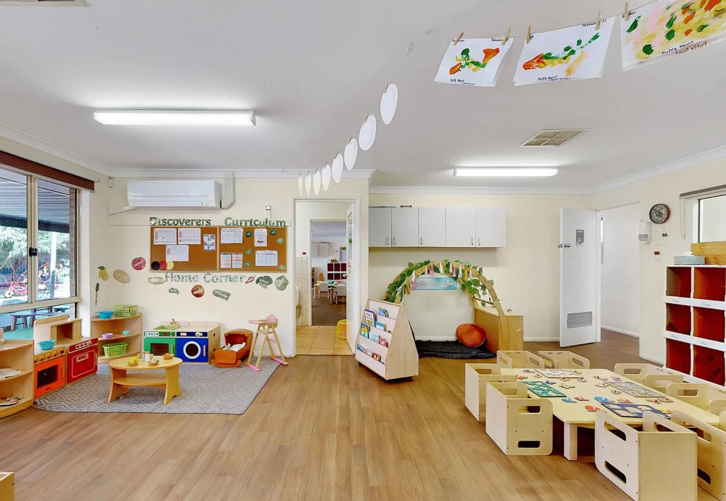 Bright and colorful preschool classroom with labeled activity areas, child-sized furniture, and art projects hanging from a string.