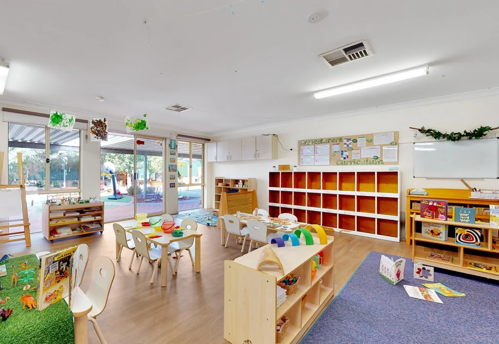 A brightly lit classroom with child-sized chairs and tables, bookshelves, educational toys, and learning materials. There are windows showing an outdoor play area.